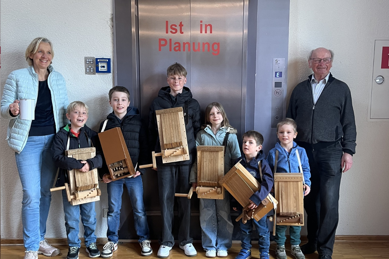 Das Bild zeigt eine Gruppe von acht Personen, darunter Kinder und Erwachsene, die vor einer Aufzugtür stehen. Die Kinder halten selbstgebaute Holzratschen in den Händen. Auf der Tür steht "1st in Planung". Alle lächeln und posieren für das Foto.