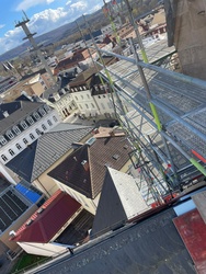 Das Bild zeigt eine Aussicht aus großer Höhe auf eine Stadt mit Dächern und einem Hügel im Hintergrund. Vordergrund: Ein Baugerüst an einer Kirchturmspitze, daneben ein rotes Dach.