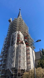 Das Bild zeigt einen hohen Kirchturm, der mit einem Baugerüst umgeben ist. Das Gerüst bedeckt den Turm komplett, und oben befindet sich eine Turmspitze mit einem Wetterhahn. Der Himmel ist klar und blau.