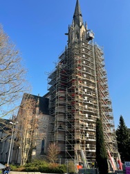 Sanierung Turm Herz Jesu_2026_Das Bild zeigt eine Kirche mit einem hohen, spitzen Turm, der von einem Gerüst umgeben ist. Die Kirche hat gotische Architekturmerkmale und befindet sich in einer baumbestandenen Umgebung bei klarem, blauem Himmel.