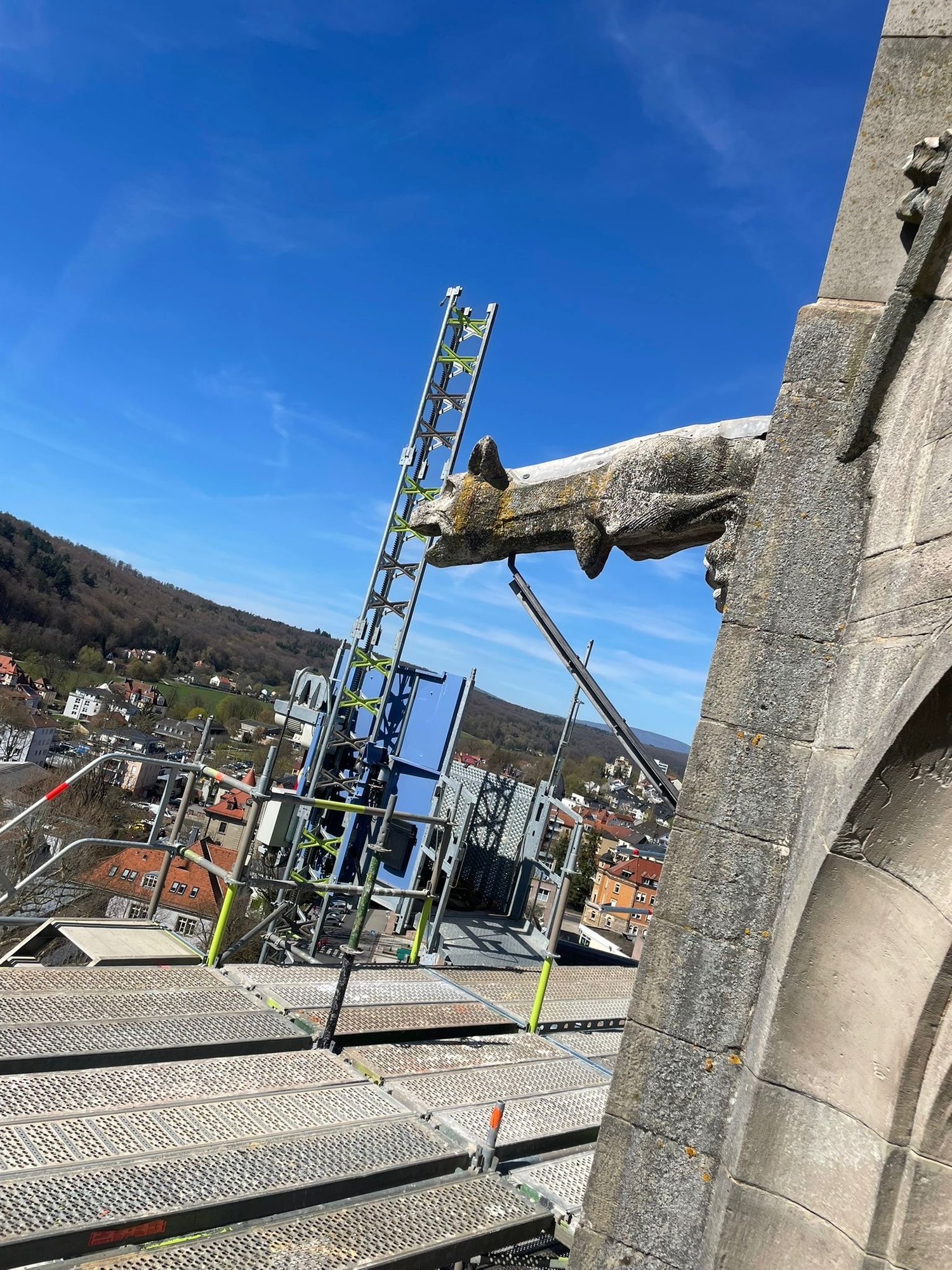 Das Bild zeigt eine Baustelle auf dem Dach einer Kirche. Man sieht Gerüste und Leitern, die an der Fassade befestigt sind. Im Hintergrund sind Häuser und Berge unter einem klaren blauen Himmel zu sehen.