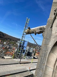 Das Bild zeigt eine Baustelle auf dem Dach einer Kirche. Man sieht Gerüste und Leitern, die an der Fassade befestigt sind. Im Hintergrund sind Häuser und Berge unter einem klaren blauen Himmel zu sehen.