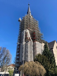Das Bild zeigt eine große Kirche mit einem hohen Turm, der von einem umfangreichen Baugerüst umgeben ist. Der Turm hat eine spitze Fassade mit einem Kreuz obenauf. Der Himmel ist klar blau, und es gibt einige Bäume und Gebäude im Hintergrund.