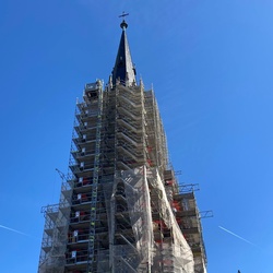 Das Bild zeigt einen Kirchturm, der mit Baugerüsten umgeben ist. Die Spitze des Turms ragt in den klaren blauen Himmel. Am oberen Ende befindet sich ein Kreuz.