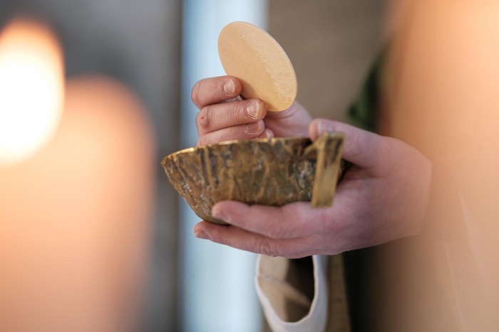 Das Bild zeigt eine Hand, die eine dünne, runde Hostie hält und eine goldene Schale mit Reliefs darauf. Die Szene wirkt feierlich und religiös.