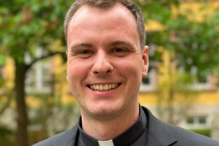 Ein Mann in einem dunklen Anzug mit einem weißen Kragen steht vor grünen Bäumen und einem Gebäude im Hintergrund. Er lächelt und schaut direkt in die Kamera.