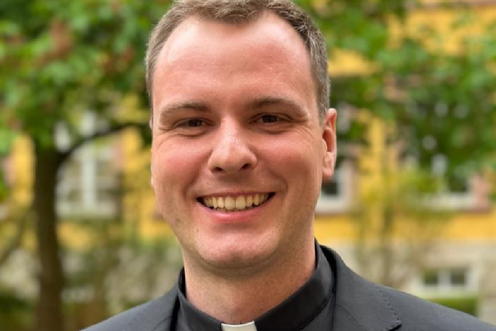 Ein Mann in einem dunklen Anzug mit einem weißen Kragen steht vor grünen Bäumen und einem Gebäude im Hintergrund. Er lächelt und schaut direkt in die Kamera.